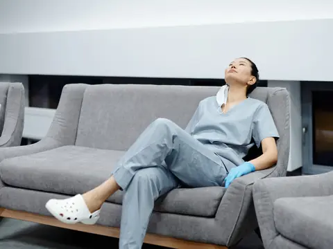 A nurse in scrubs and clogs resting on a couch after a shift